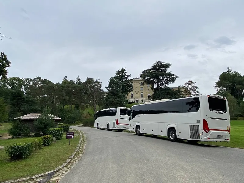 Bus hire in Paris offering with view of two elegant white buses ready for group travel, parked on a scenic tree-lined road beside a quaint gazebo, with a classic French chateau in the background.
