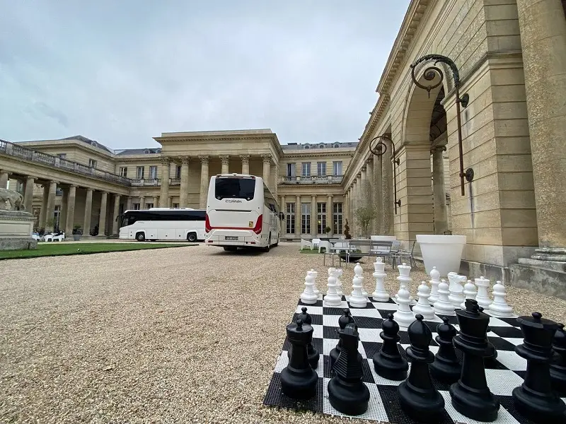 Bus charter Paris service highlighted by two pristine white buses positioned near a majestic neoclassical building, complemented by an oversized chess set in the foreground.
